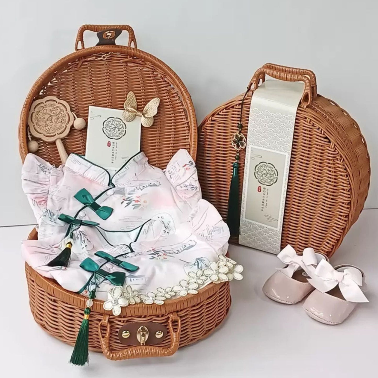 A baby gift set including a onesie, romper, headband, socks, and shoes, presented in a wicker basket with a rattle and a gift bag.