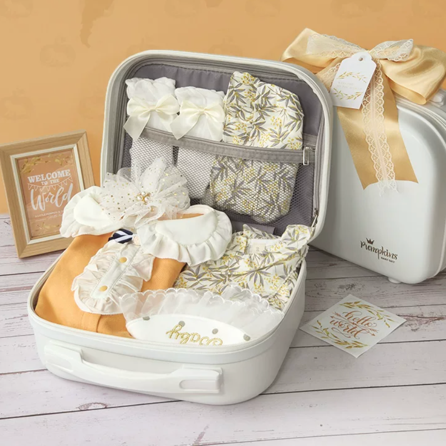 A baby gift set arranged inside an open white suitcase, with matching cream and gold patterned fabric pieces, lace bows, and a small framed sign on a light wood surface. A branded cream suitcase with a large ribbon bow sits beside it, and a card reading 'welcome to the wild' is placed nearby.